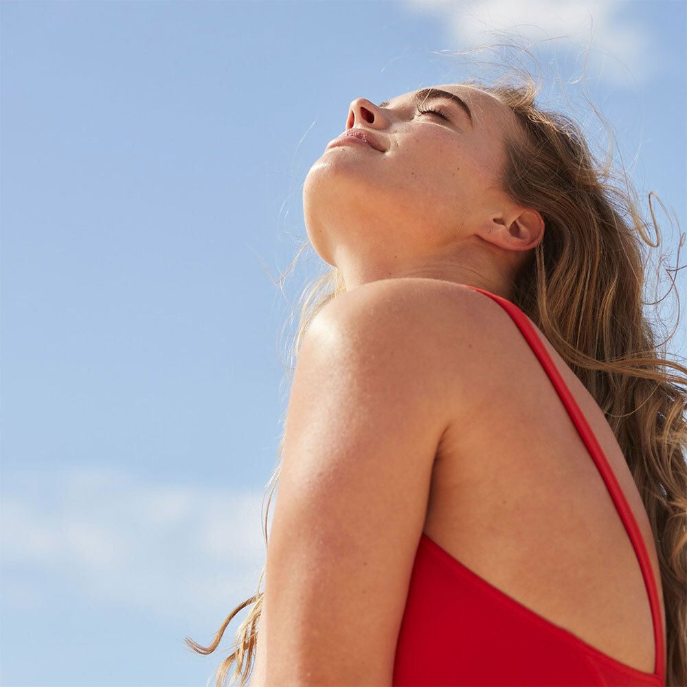 Eine Frau in einem roten Badeanzug, die ihren Kopf in den Himmel neigt. Ihr Haar ist wellig.