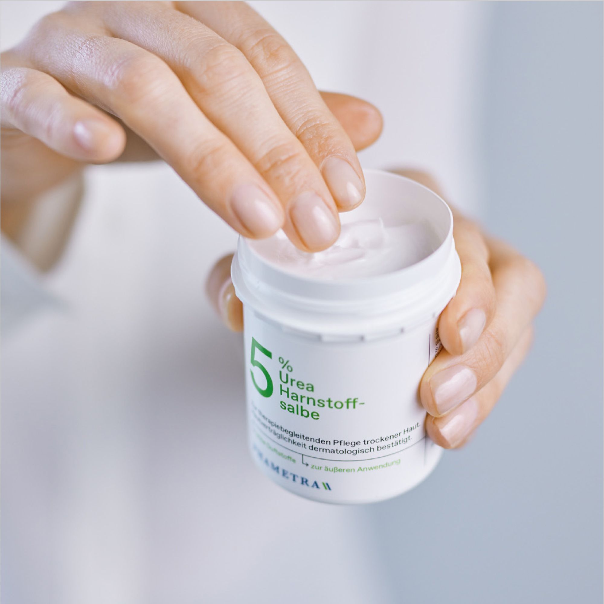 Hände öffnen eine weiße Dose mit Creme. Auf der Dose steht: 5% Urea Harnstoffsalbe. Marke: PHARMETRA.