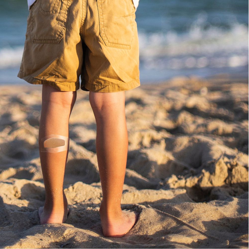 Ein Kind am Strand mit einem Pflaster am Bein. Pflaster ist transparent.