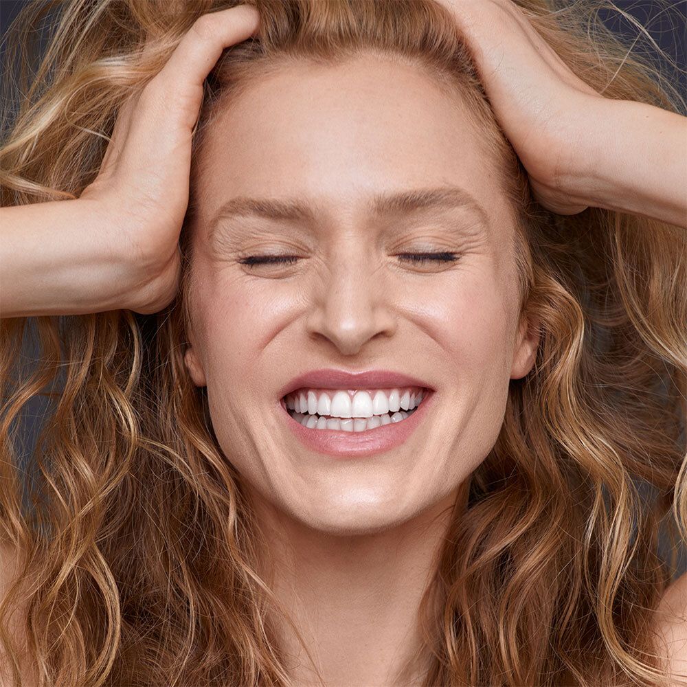 Frau mit blonden Locken, Hände im Haar, Augen geschlossen, lächelnd.