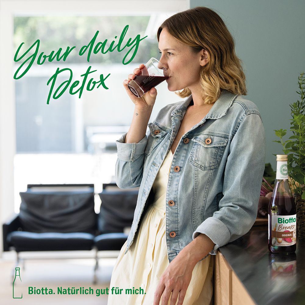Frau trinkt Saft aus Glas. Flasche Biotta Breuss Antioxidant im Hintergrund. Text: Your daily Detox.