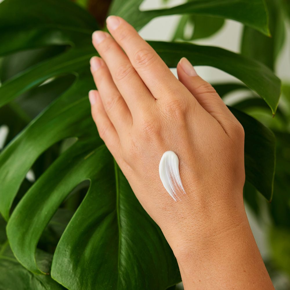 Hand mit Creme auf der Haut. Grüne Blätter im Hintergrund. Creme-Klecks sichtbar.