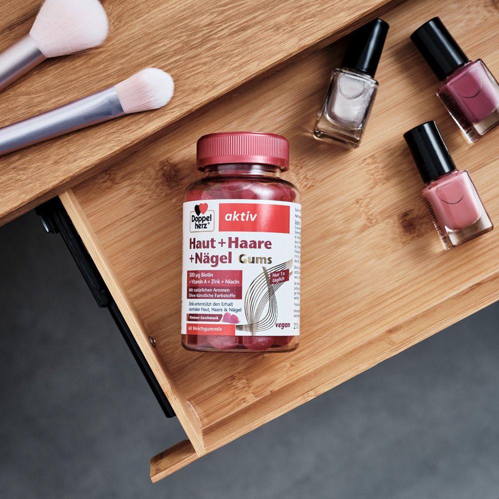 Doppelherz aktiv Haut + Haare + Nägel Gums auf Holzablage. Daneben Make-up-Pinsel und Nagellack. Rote Gummibärchen.