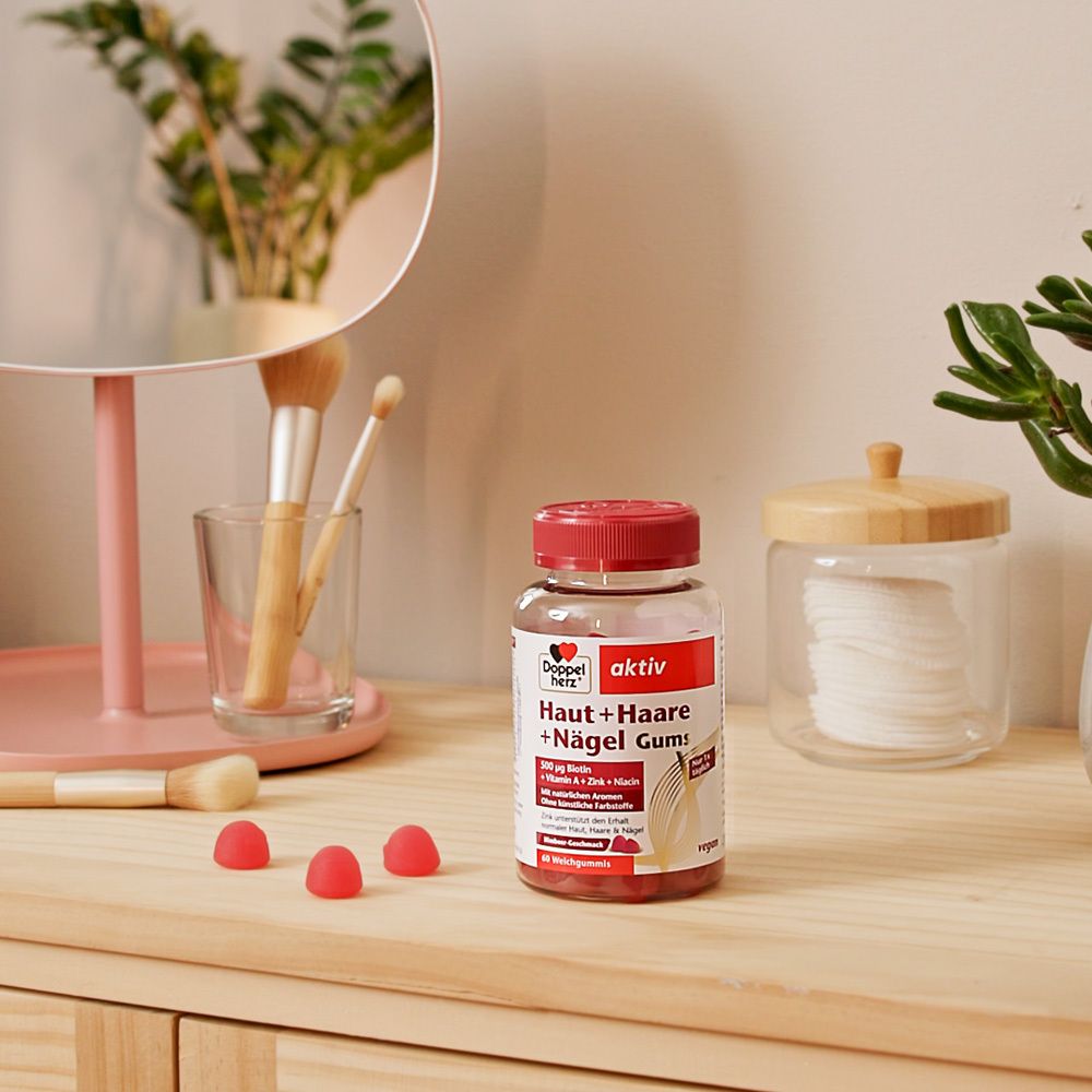Doppelherz aktiv Haut + Haare + Nägel Gums auf Holz. Daneben Make-up-Zubehör. Rote Gummibärchen. Vegan.