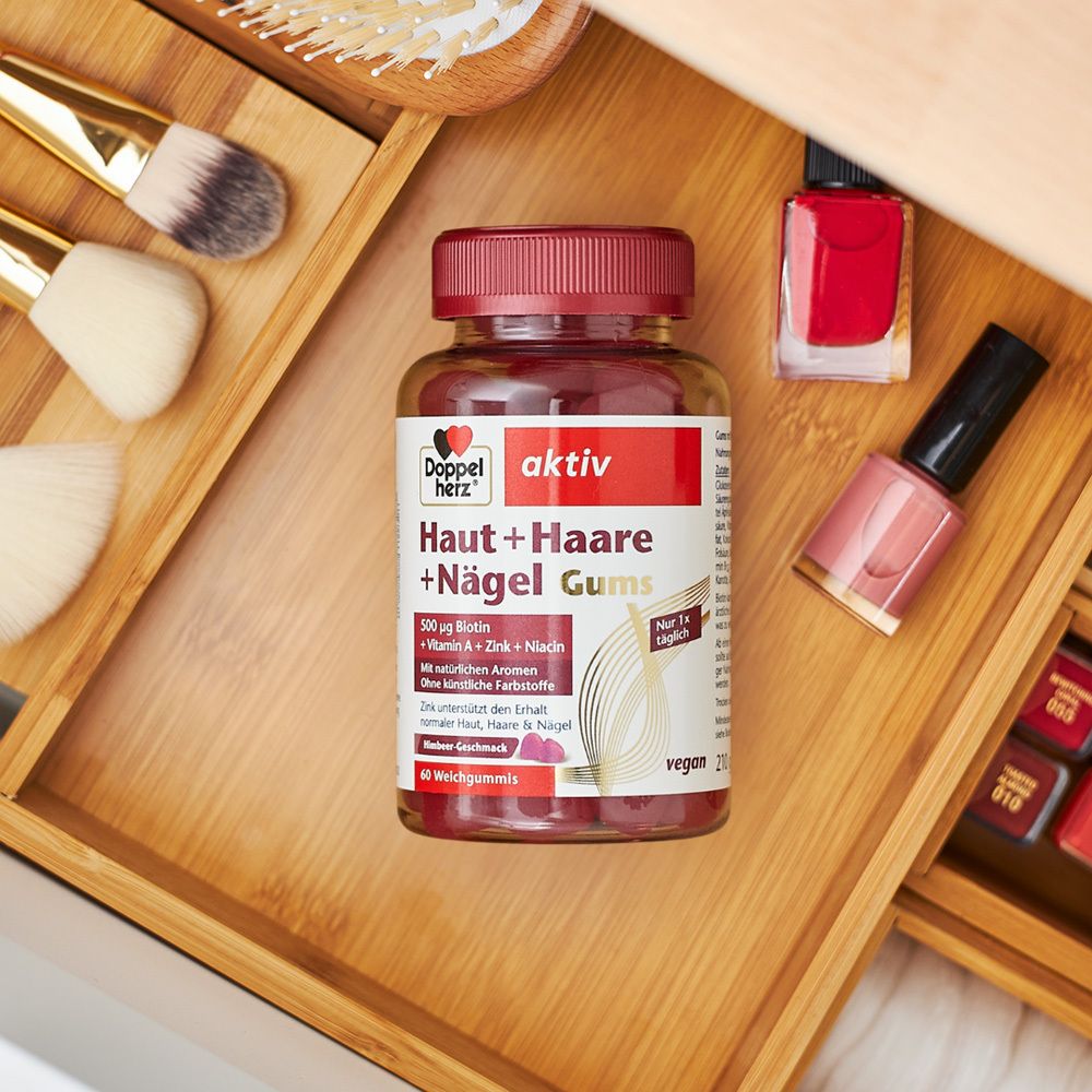 Doppelherz aktiv Haut + Haare + Nägel Gums auf Holzablage. Daneben Nagellack und Make-up-Zubehör. Rote Gummibärchen.