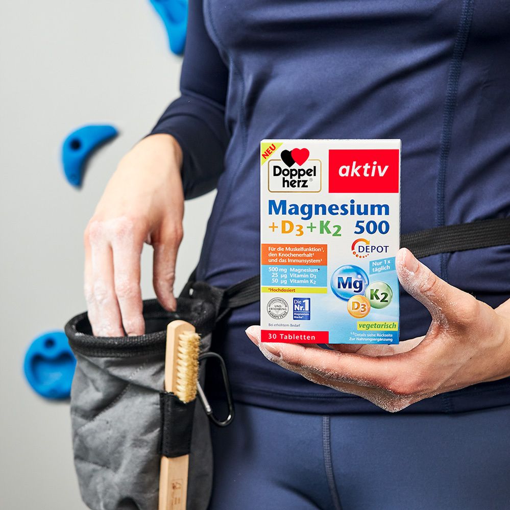 Person hält eine Packung Doppelherz® Magnesium + D3 + K2 500. Im Hintergrund Kletterwand.