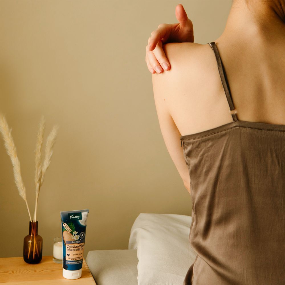 Kneipp® Gute Nacht Körpercreme neben einer Frau. Die Frau berührt ihre Schulter. Auf einem Nachttisch steht die Creme.
