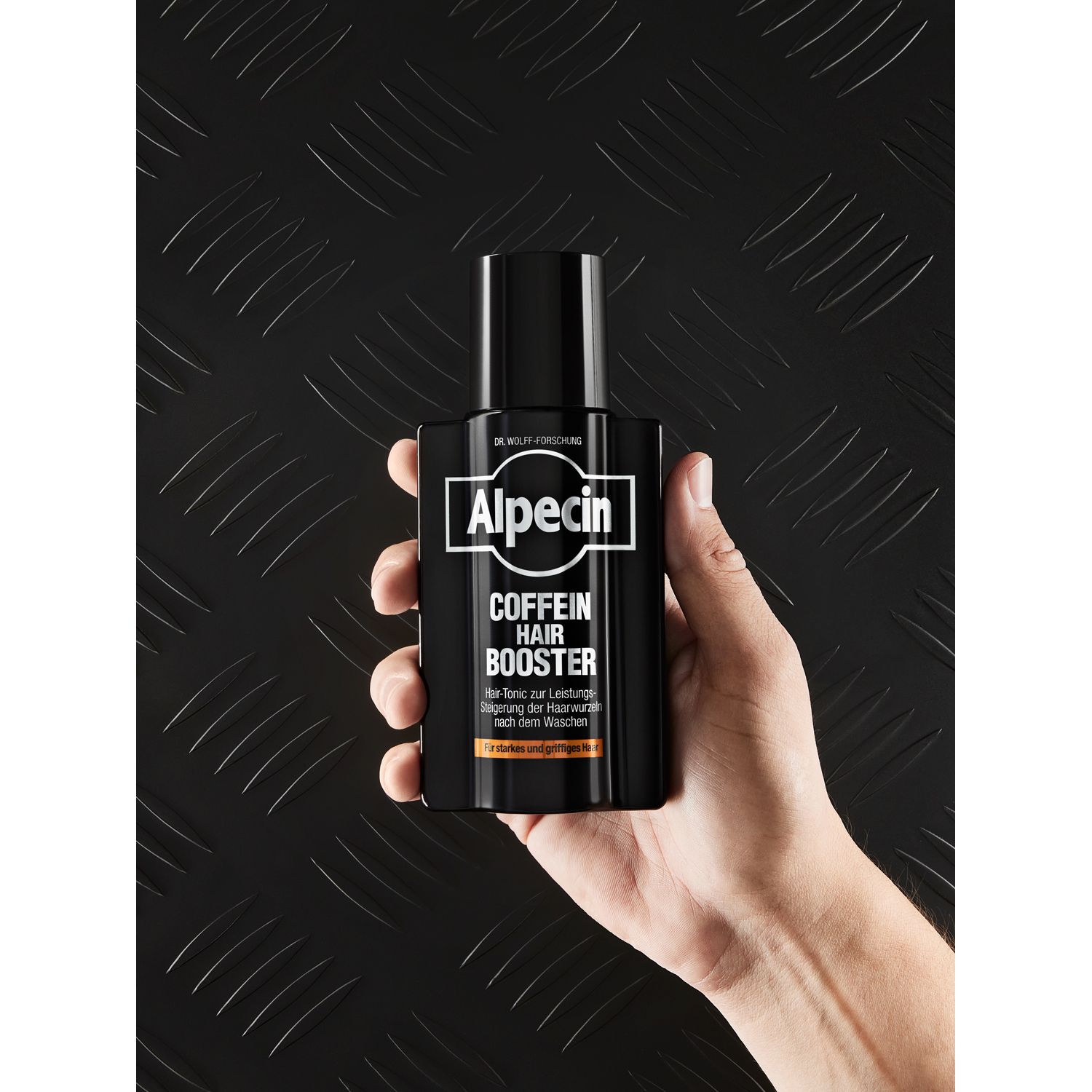 Hand hält eine schwarze Flasche mit Alpecin-Logo und 'Coffein Hair Booster'. Hintergrund: Metallstruktur.