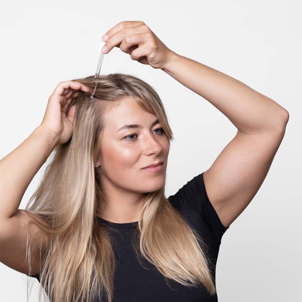 Frau mit Pipette am Haaransatz. Blondes Haar, schwarzes Oberteil.