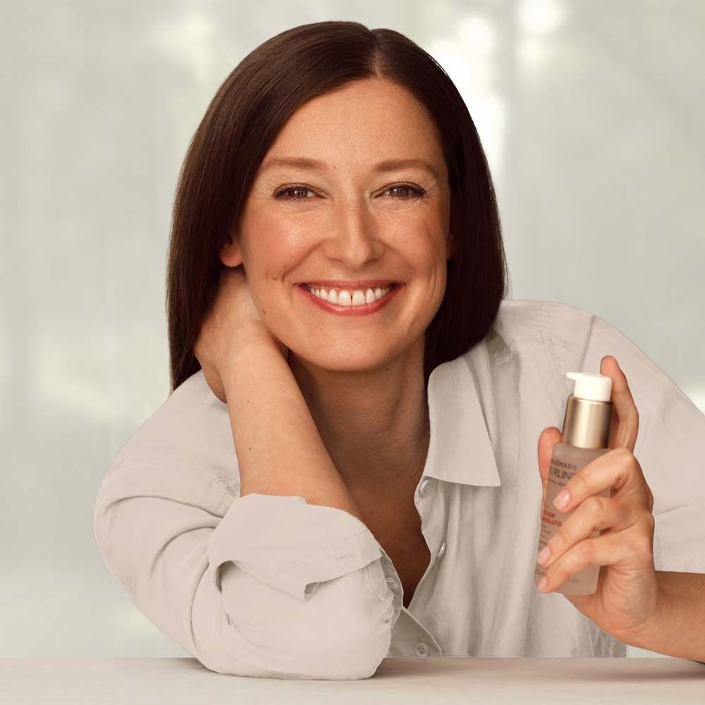 Frau hält Börlind System Absolute Beauty Fluid. Helle Kleidung. Lächelt. Produkt in der Hand.