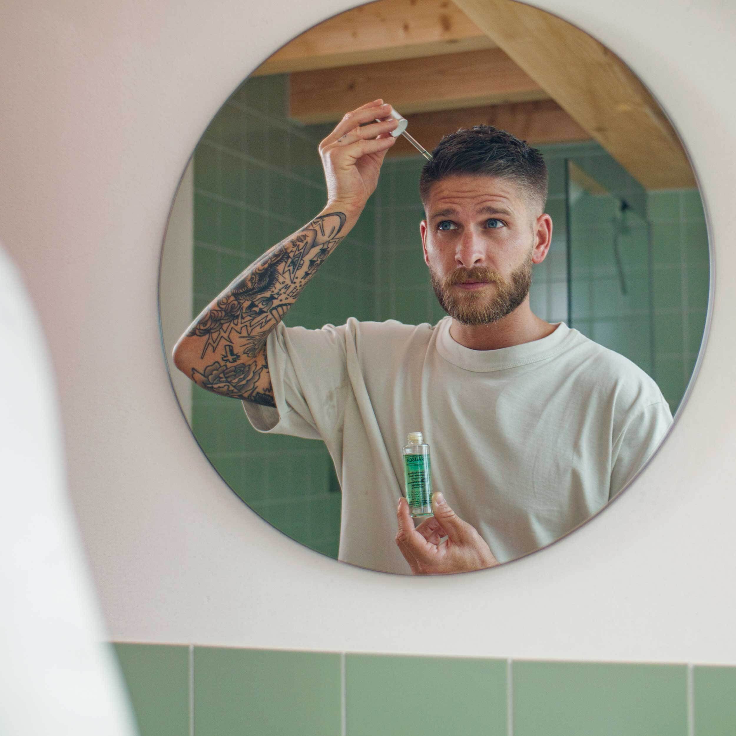 Mann mit Bart und Tattoos trägt Tropfen aus einer kleinen grünen Flasche mit Pipette auf die Kopfhaut.