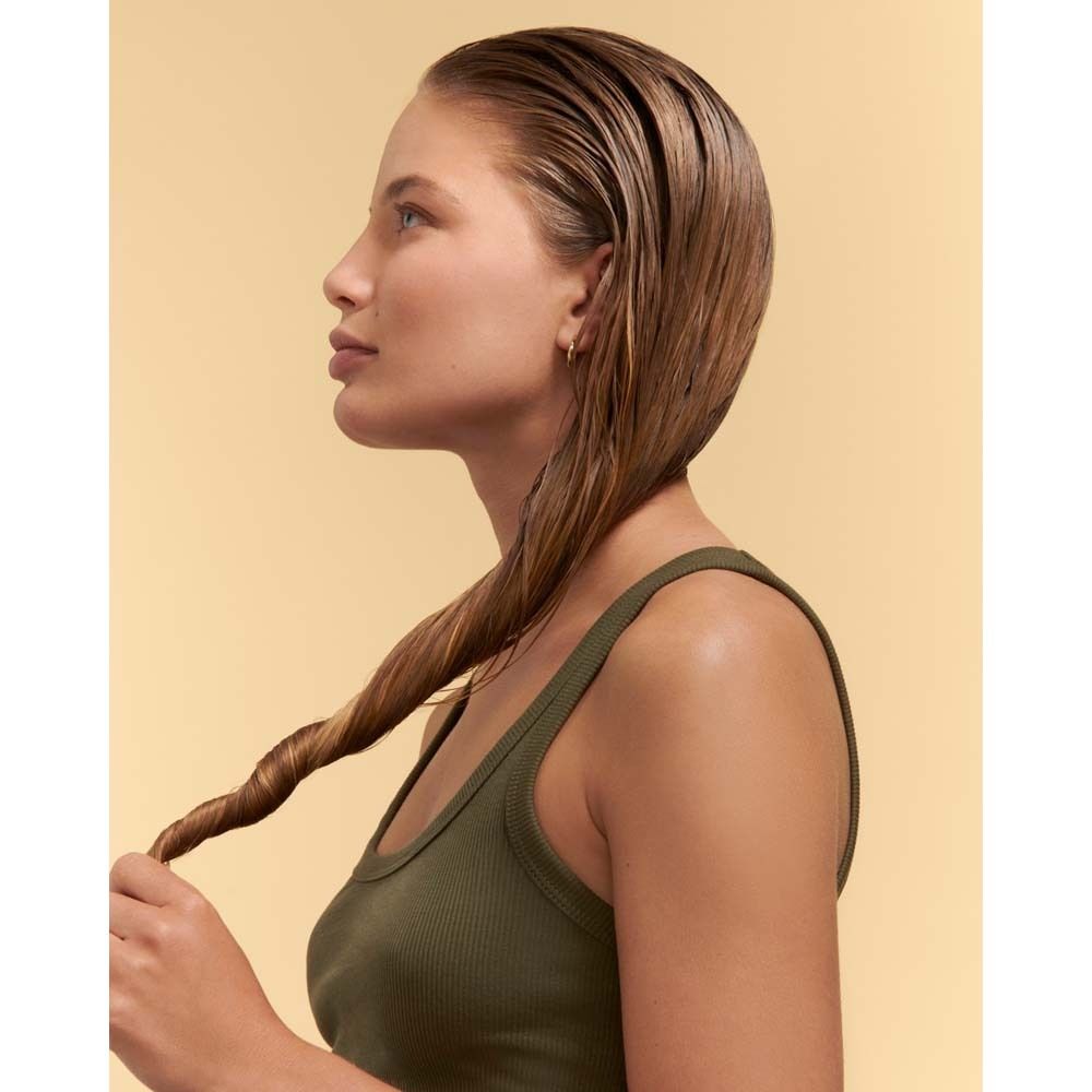 Frau mit nassem Haar, das zu einem Zopf geflochten wird. Sie trägt ein olivgrünes Tanktop. Gelber Hintergrund.