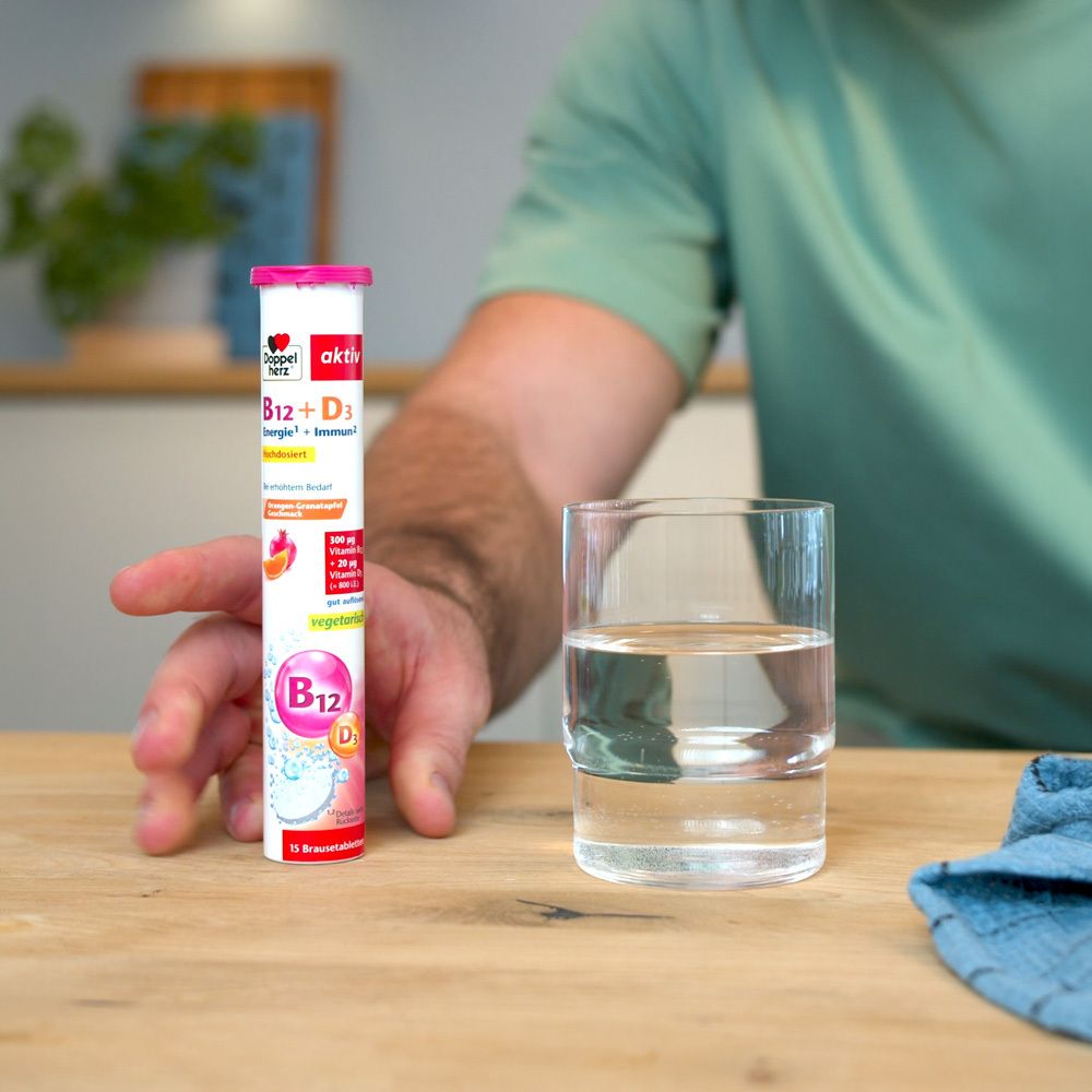 Person hält eine Tube Doppelherz aktiv B12 + D3 neben einem Glas Wasser. Tischplatte aus Holz.