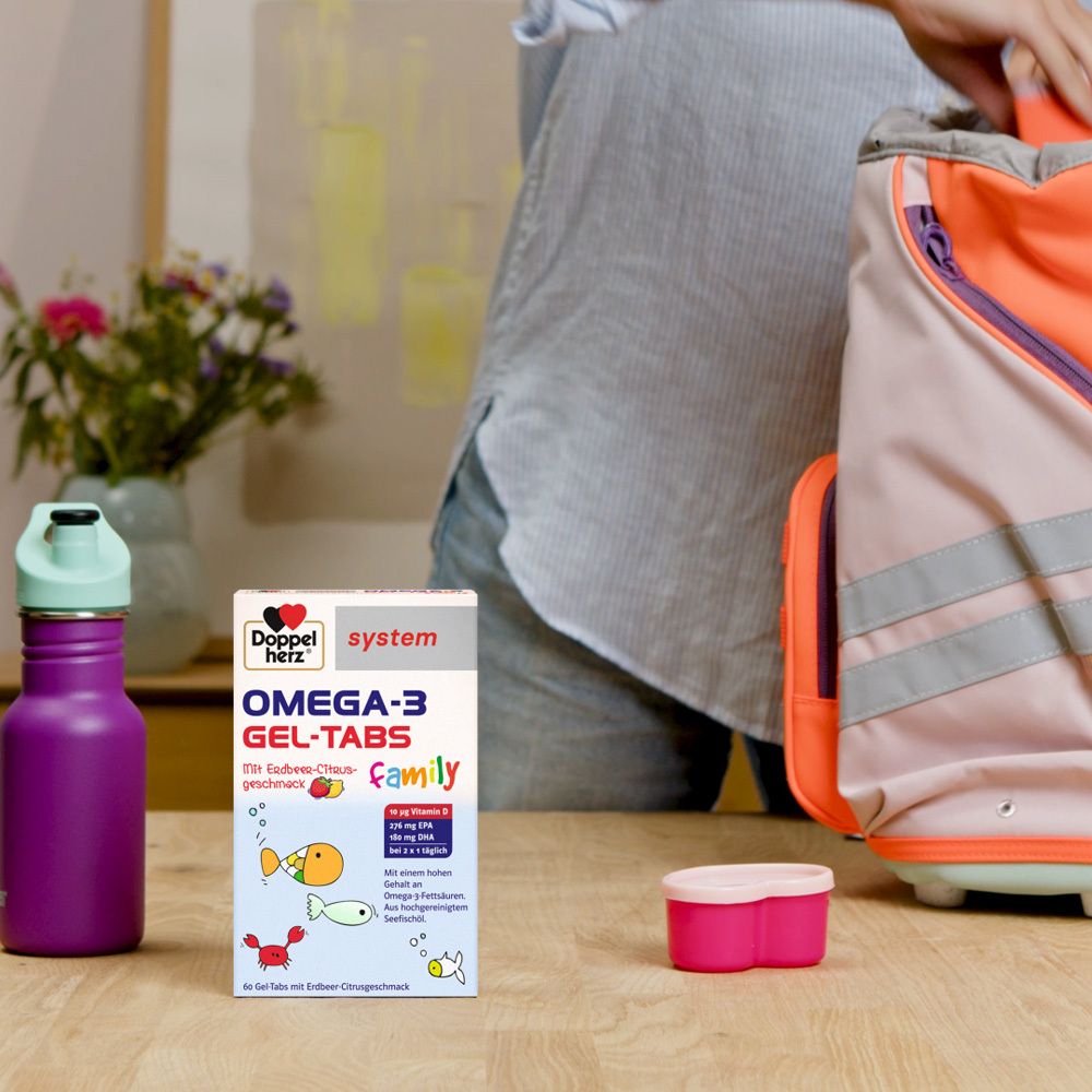Doppelherz system OMEGA-3 GEL-TABS Family Packung auf einem Tisch. Daneben eine Trinkflasche und eine Brotdose.
