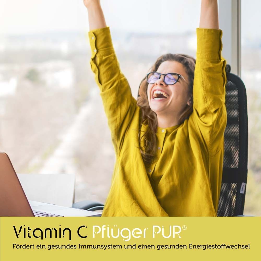 Vitamin C Pflüger® PUR. Frau mit Brille hebt Arme. Gelbes Oberteil. Büro-Hintergrund.