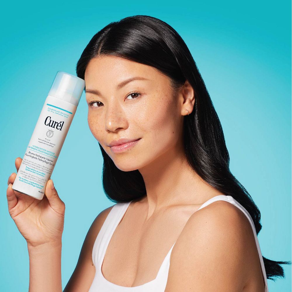 Asiatische Frau hält weißes Spray mit hellblauem Deckel. Aufschrift: Curél. Für trockene, empfindliche Haut. Hintergrund: blau.