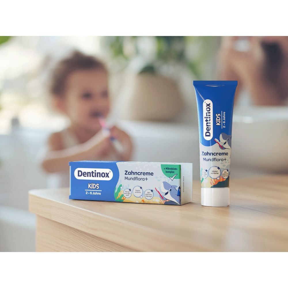 Zahncreme-Tube und -Karton neben Waschbecken. "Dentinox KIDS" und "Zahncreme Mundflora+" sichtbar. Kind putzt Zähne im Hintergrund.