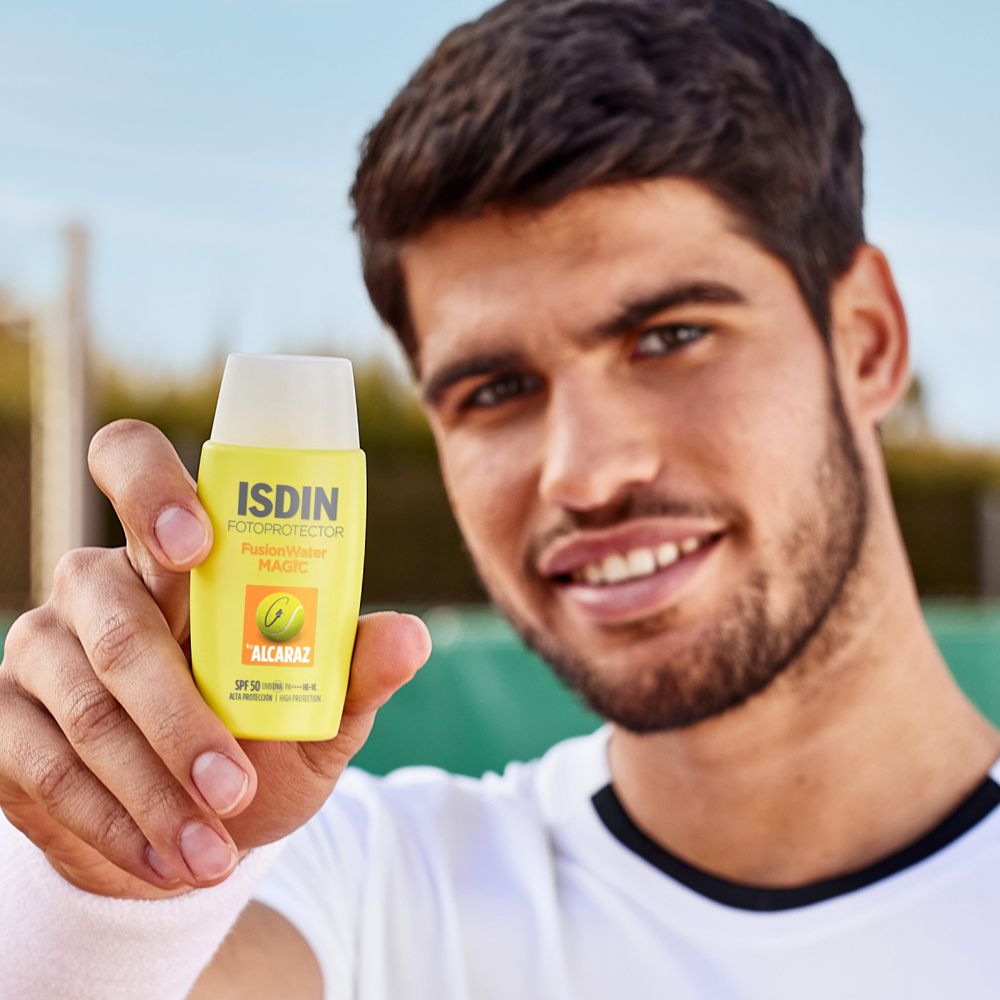 Mann hält ISDIN FusionWater MAGIC Alcaraz. Gelbe Flasche. Tennisplatz im Hintergrund.