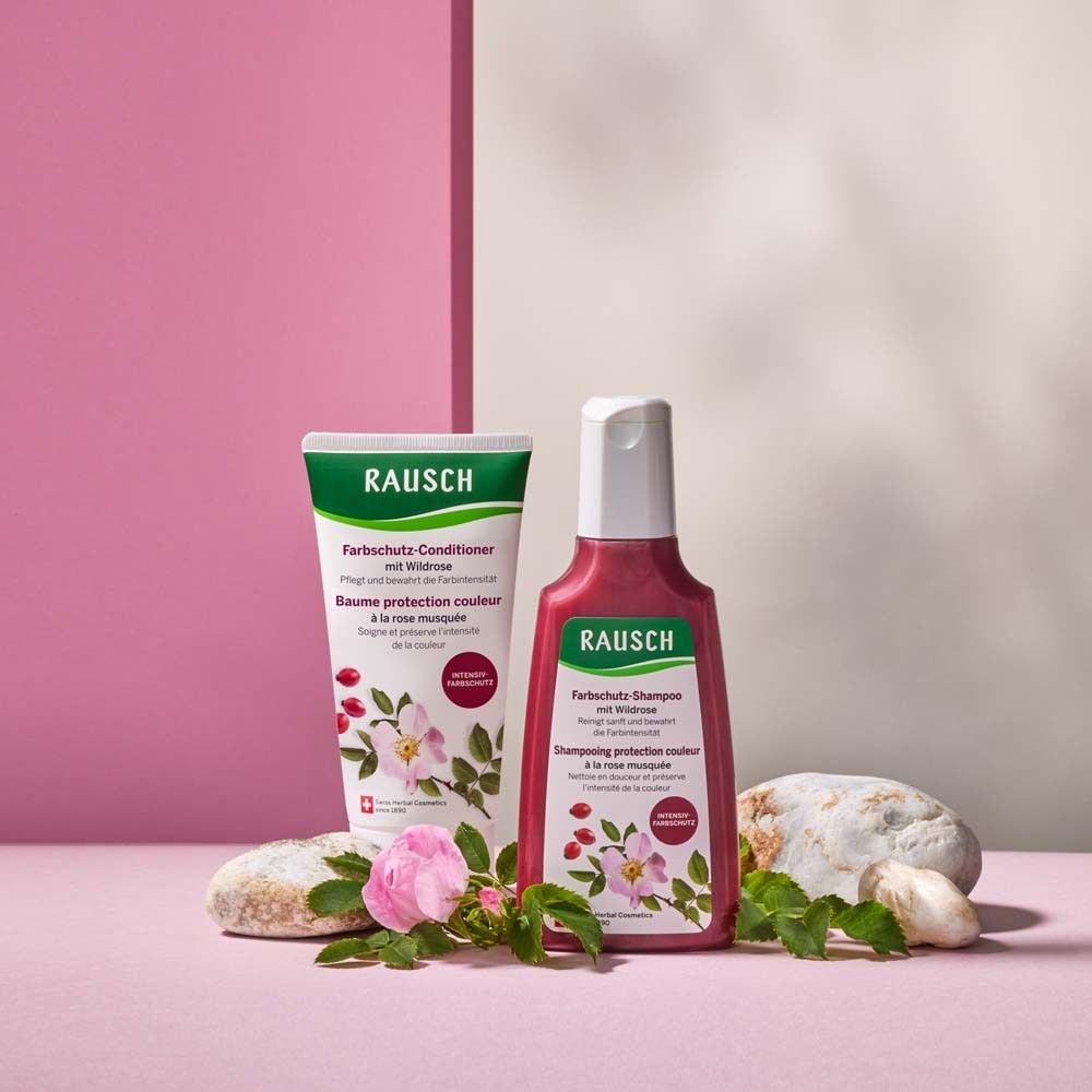 Produkt-Arrangement: Shampoo und Conditioner mit Wildrose. Auf rosa Untergrund, mit Steinen und Blüten dekoriert.