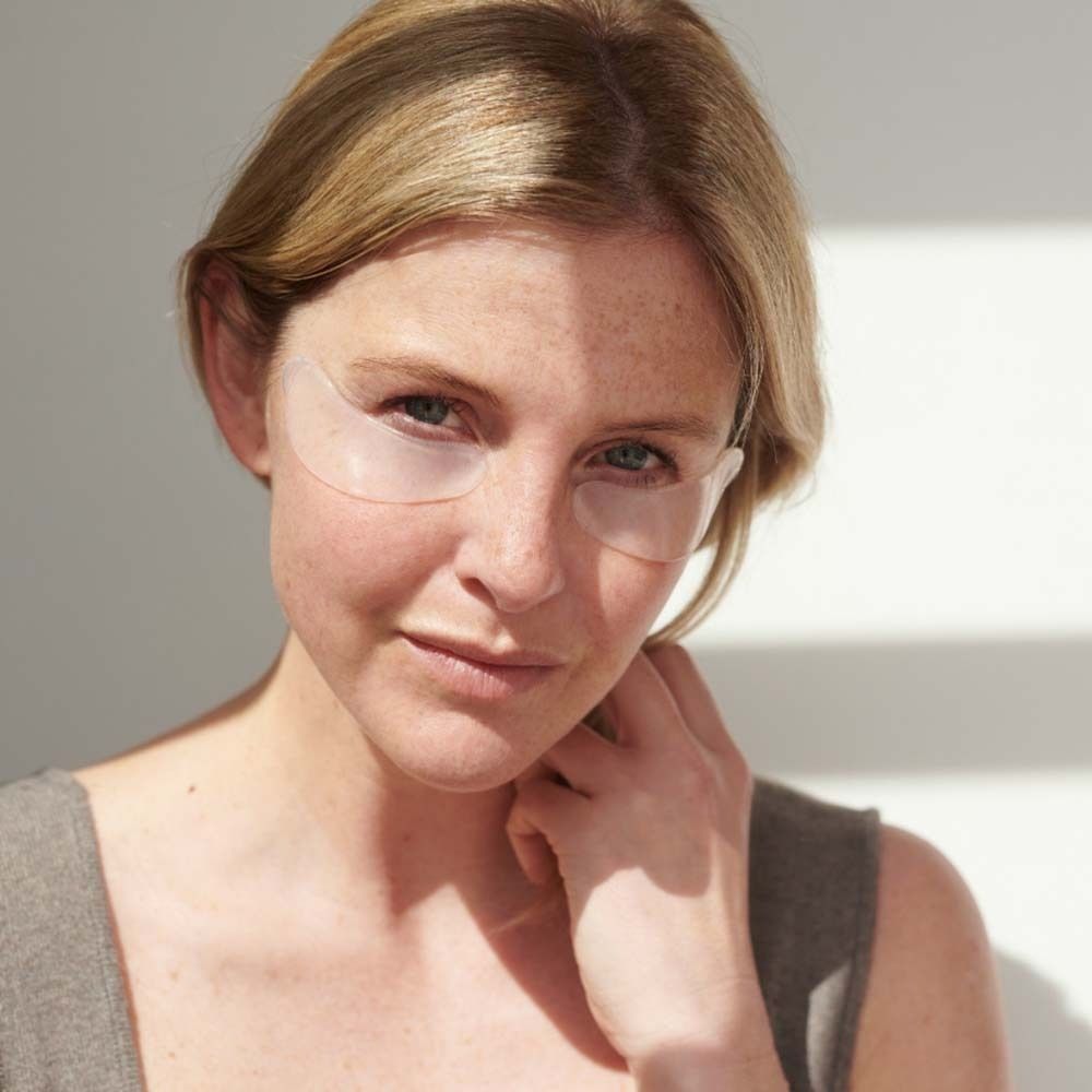 Frau mit Augen Pads unter den Augen. Sie trägt ein graues Top. Sonnenlicht im Hintergrund.