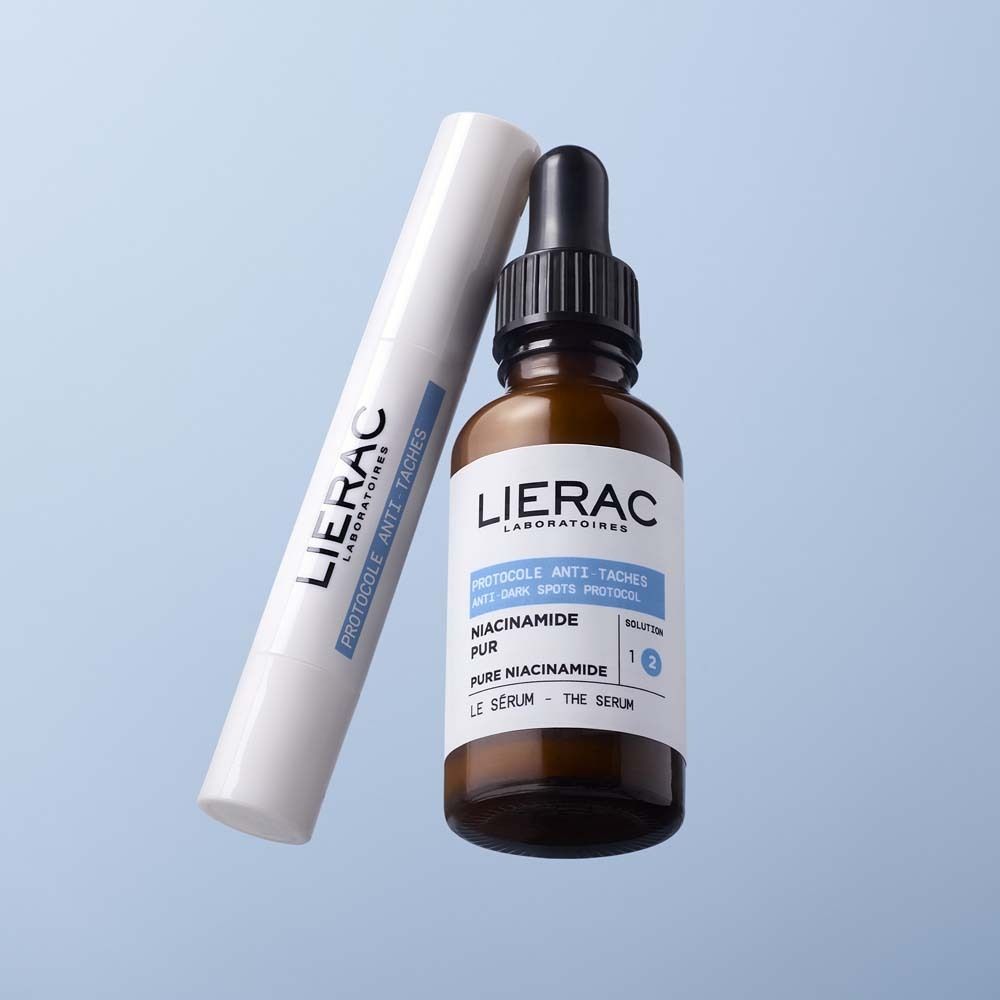 Weißer Stick und braune Flasche. LIERAC-Logo und Produktinformationen: Protocole Anti-Taches, Vitamin E, Le Stick Solution. Flasche mit Serum.