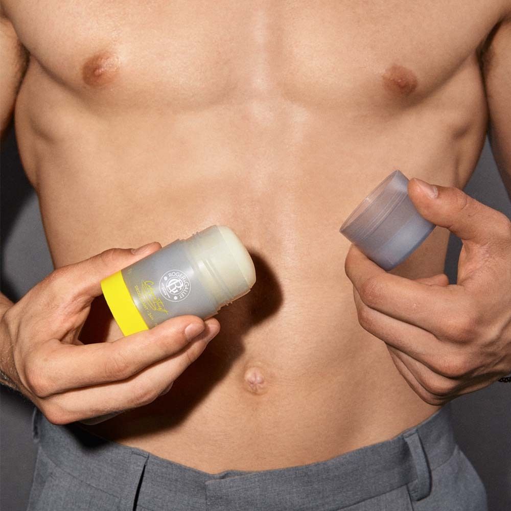 Mann hält zwei Teile eines Deodorant-Sticks. Gelbe Basis, grauer Deckel. Sichtbarer Oberkörper.