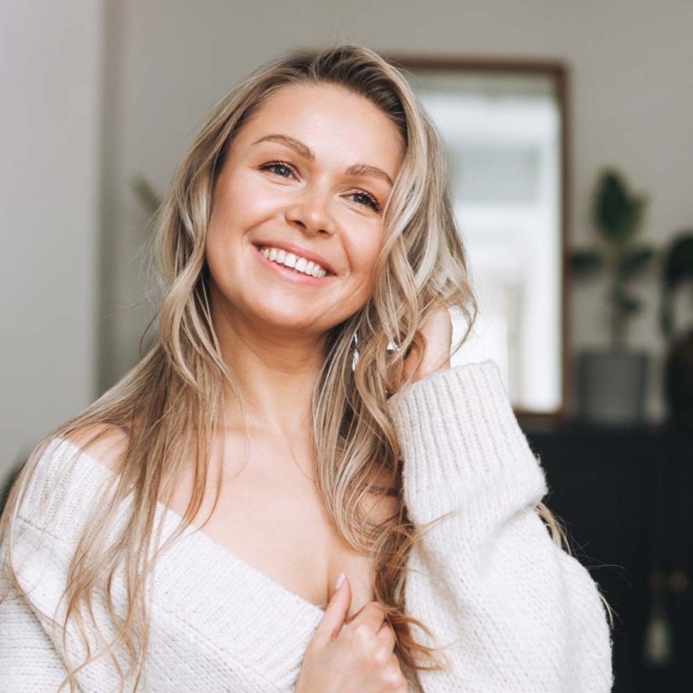 Porträt einer lächelnden blonden Frau mit langen Haaren. Sie trägt einen weißen Pullover. Der Hintergrund ist unscharf.
