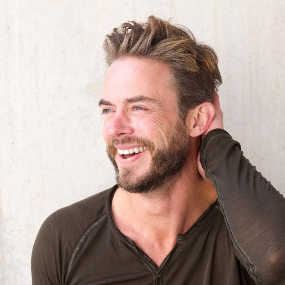 Ein lächelnder Mann mit Bart und blonden Haaren. Er berührt seinen Hinterkopf. Der Hintergrund ist hellgrau.