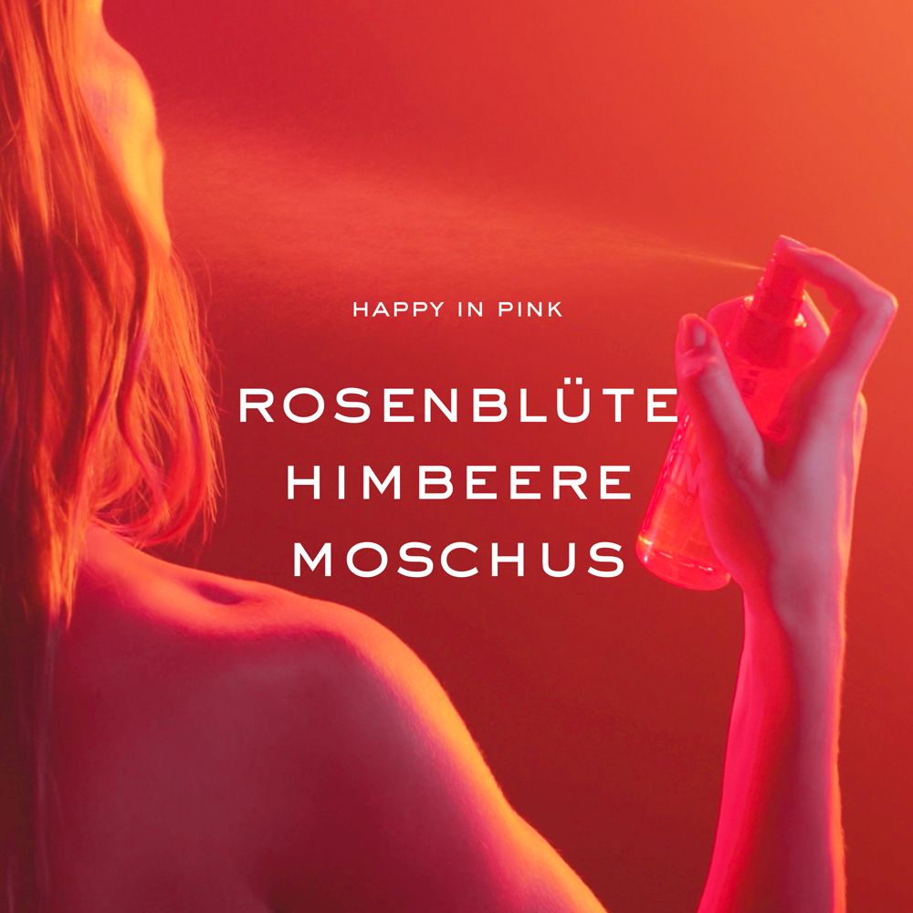 Person sprüht NUXE® Happy In Pink. Aufschrift: Rosenblüte, Himbeere, Moschus. Roter Hintergrund. Rosa Sprühnebel.
