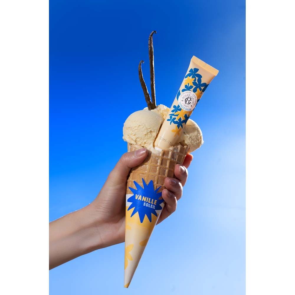 Handcreme-Tube in Eistüte mit Eis und Vanilleschoten. Hintergrund: blauer Himmel. Hand hält die Eistüte.