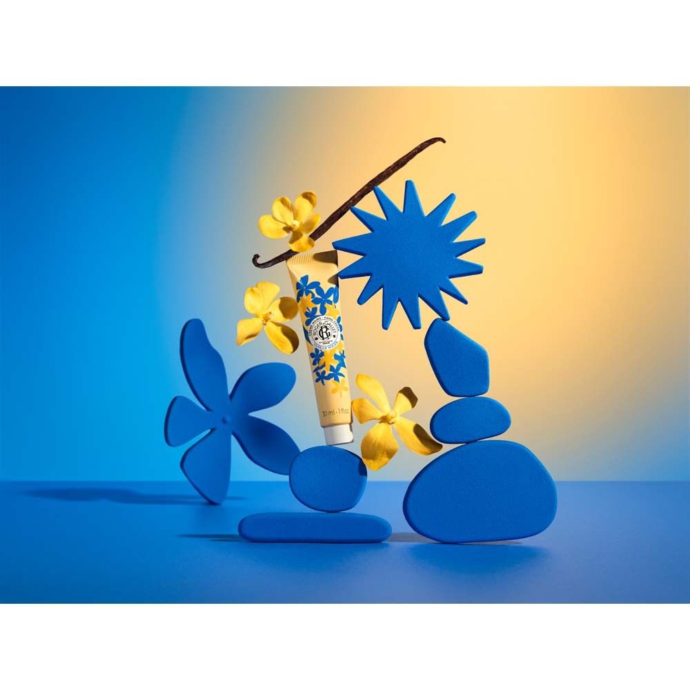 Arrangement mit Handcreme-Tube, blauen und gelben Formen, Vanilleschote und Blüten. Auf blauem Grund.