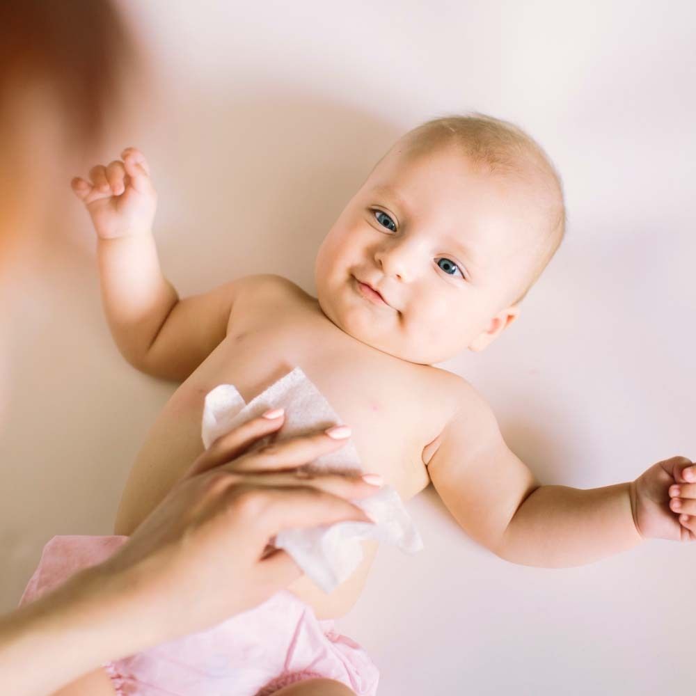 Baby liegt auf dem Rücken, eine Hand reinigt den Oberkörper mit einem weißen Tuch. Rosa Windel.