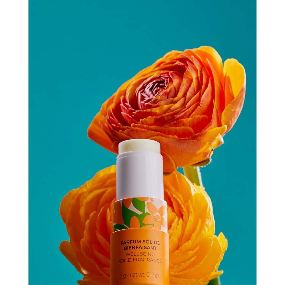 Parfumstick vor orangefarbenen Blüten. Weißer Deckel, orangefarbener Boden. Text: PARFUM SOLIDE BIENFAISANT.