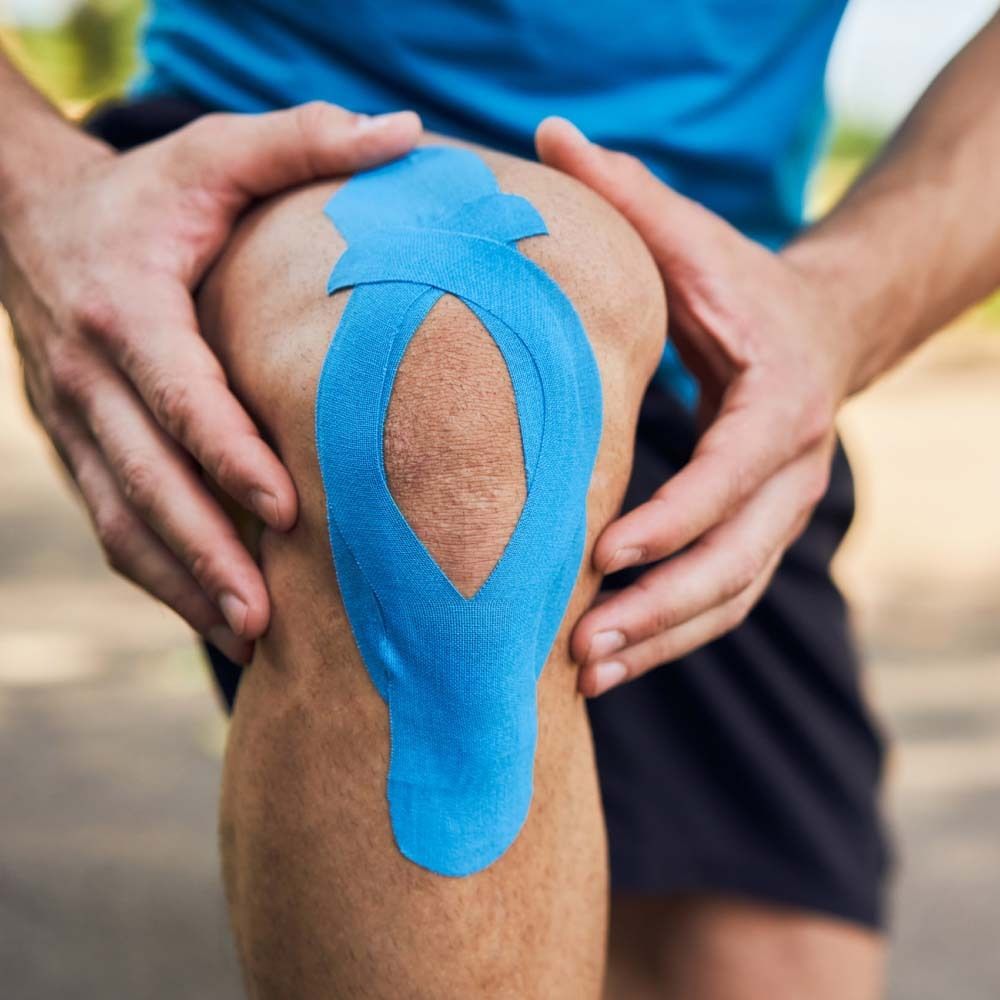 Knie mit blauem Kinesiologie Tape beklebt. Hände halten das Knie.