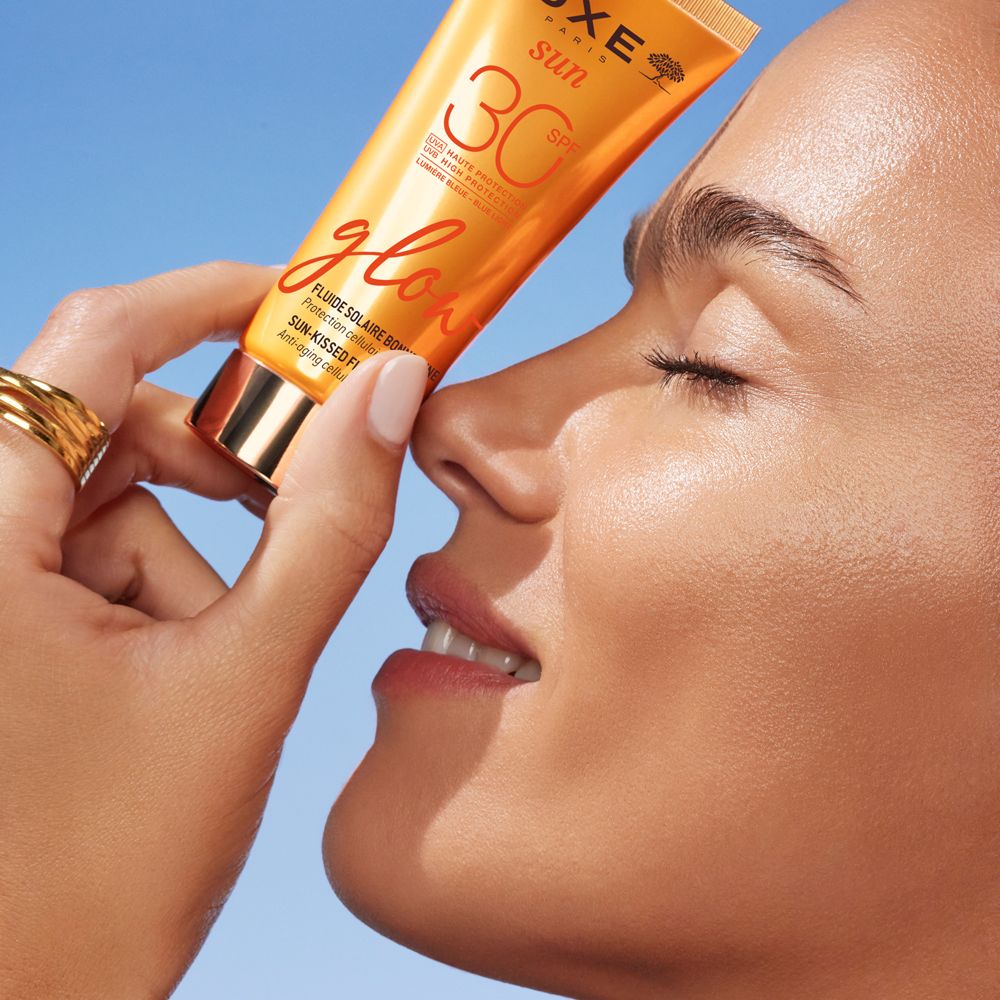 Frau hält Tube NUXE Sun Sonnenfluid Glow LSF 30. Text: SPF 30, glow, Fluide Solaire Bonne Mine.