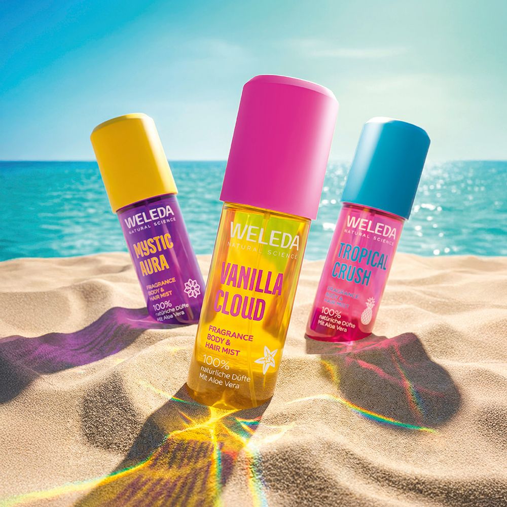 Drei Weleda Flaschen auf Sand. Mystic Aura, Vanilla Cloud, Tropical Crush. Gelb, pink, blau.