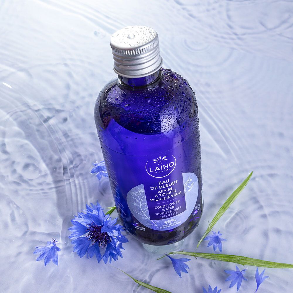 Blaue Flasche LAINO Kornblumenwasser in Wasser. Blaue Kornblumen und grüne Blätter umgeben die Flasche. Silberner Deckel.