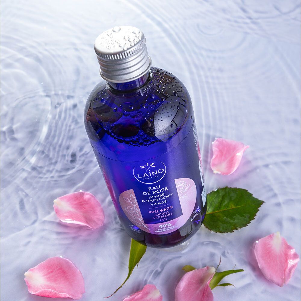 Blaue Flasche mit silbernem Deckel in Wasser mit Rosenblättern. Aufschrift: LAINO, Eau de Rose, beruhigt & erfrischt das Gesicht.
