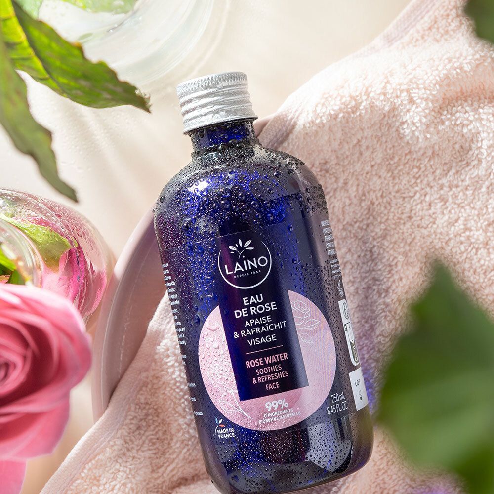 Blaue Flasche mit silbernem Deckel auf Handtuch. Aufschrift: LAINO, Eau de Rose, beruhigt & erfrischt das Gesicht. Rosen und Glas.