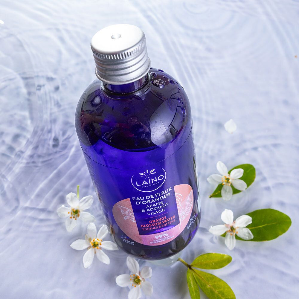 Blaue Glasflasche mit silbernem Deckel, LAINO Orangenblütenwasser, umgeben von Blüten und Wasser. Text: Eau de Fleur d'Oranger.