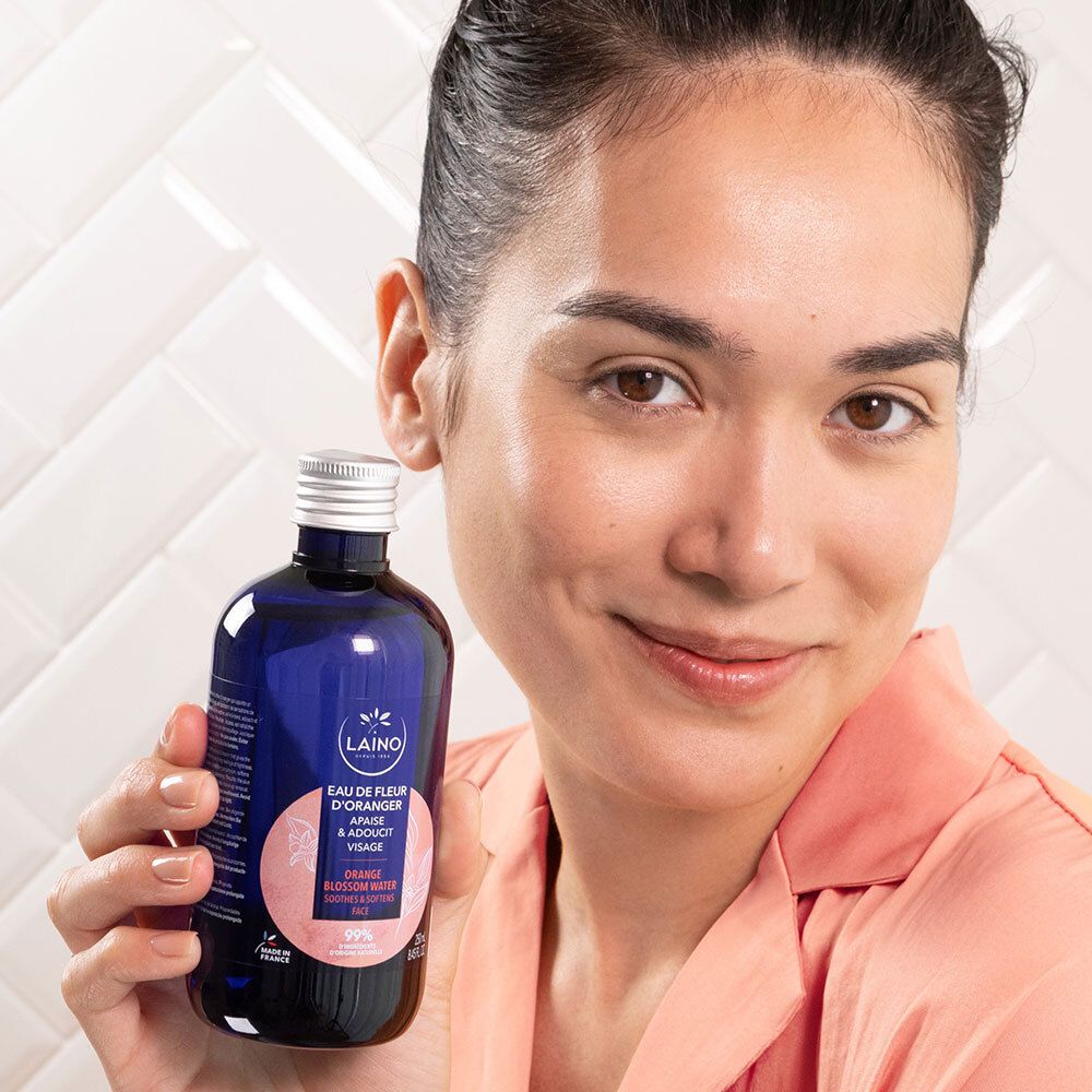 Frau hält blaue Glasflasche mit silbernem Deckel, LAINO Orangenblütenwasser. Text: Eau de Fleur d'Oranger. Frau lächelt.