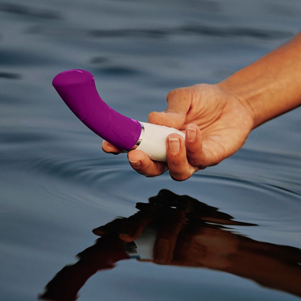 Hand hält Vibrator im Wasser. Lila-weiße Farbe. Gebogene Form.