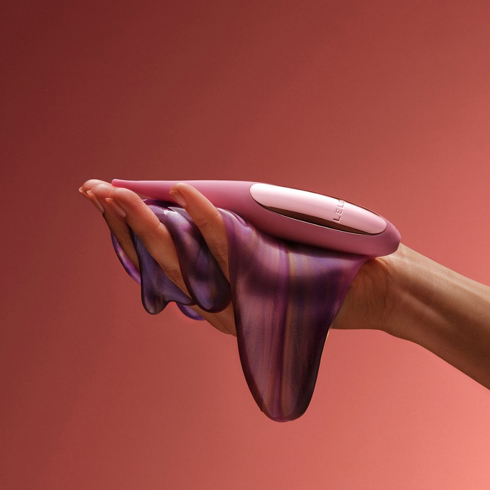 Hand hält Vibrator und eine fließende, farbige Substanz. Vibrator rosa, Marke LELO.