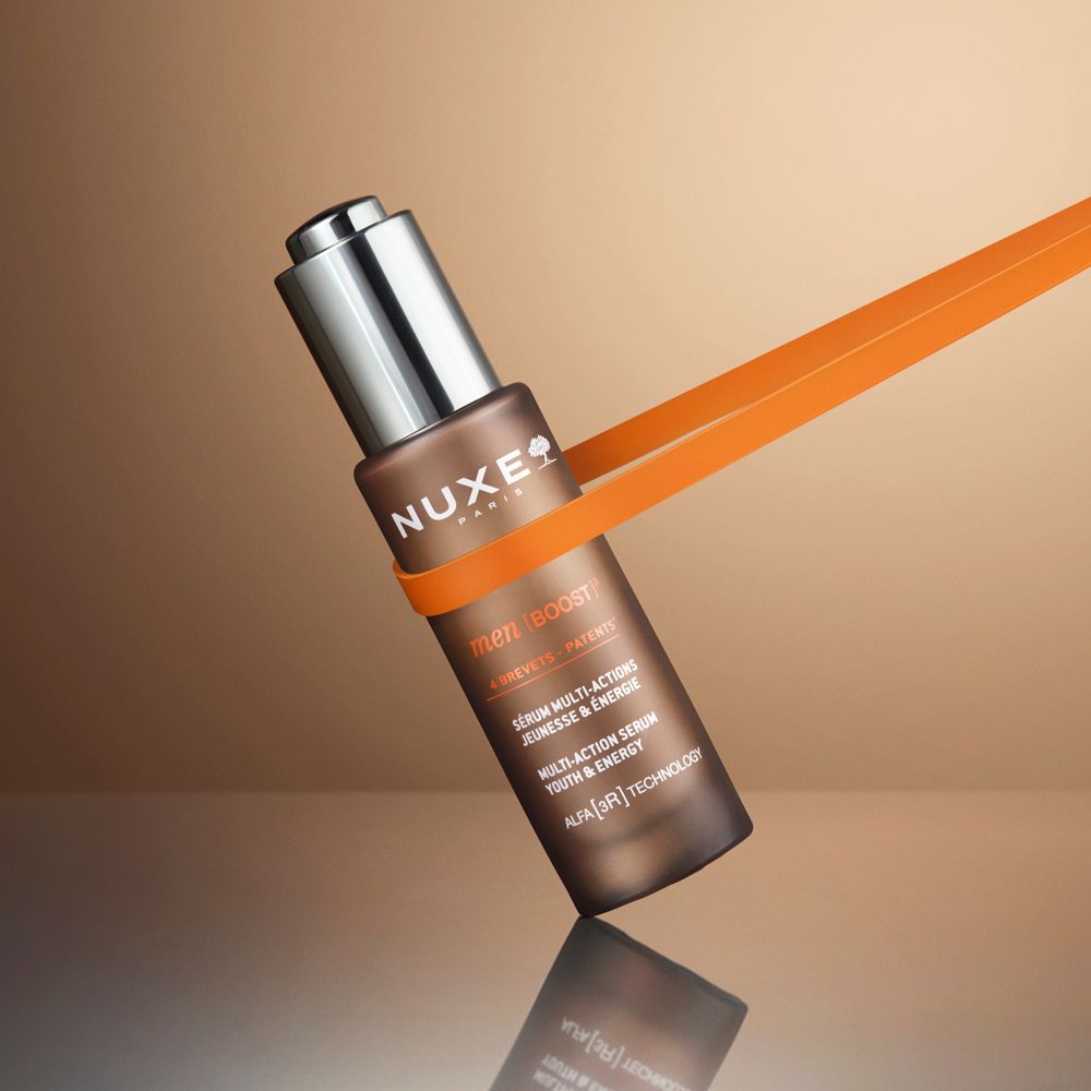 Braune Glasflasche mit silbernem Deckel, gehalten von einem orangefarbenen Band. Aufschrift: Nuxe Paris, Men Boost.