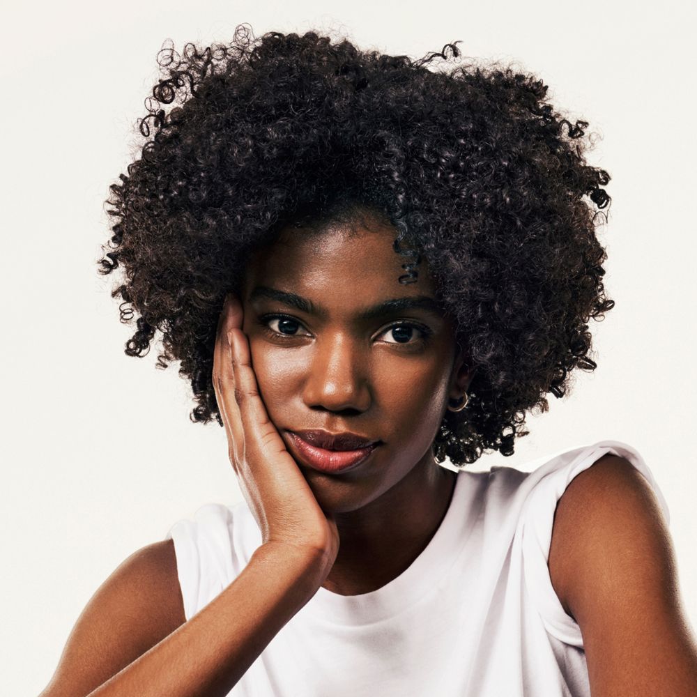 Porträt einer Frau mit lockigem Haar. Sie berührt ihre Wange. Helle Haut, weißes Oberteil.