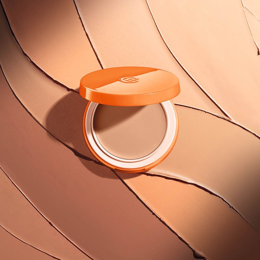 Offene, orangefarbene Kompaktpuder-Dose mit braunem Puder. Im Hintergrund Farbtöne.