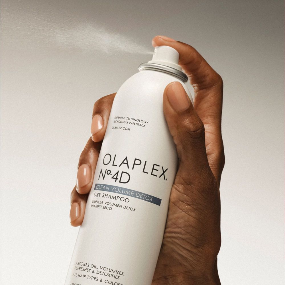 Hand hält Olaplex N°.4D Trockenshampoo-Spray. Sprühnebel ist sichtbar. Produktname und Details sind aufgedruckt.
