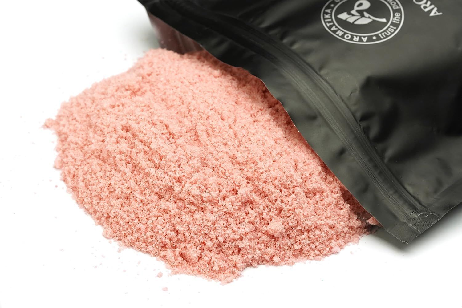 Rosa Fußbadesalz liegt aus einer geöffneten schwarzen Verpackung. Der Hintergrund ist weiß.