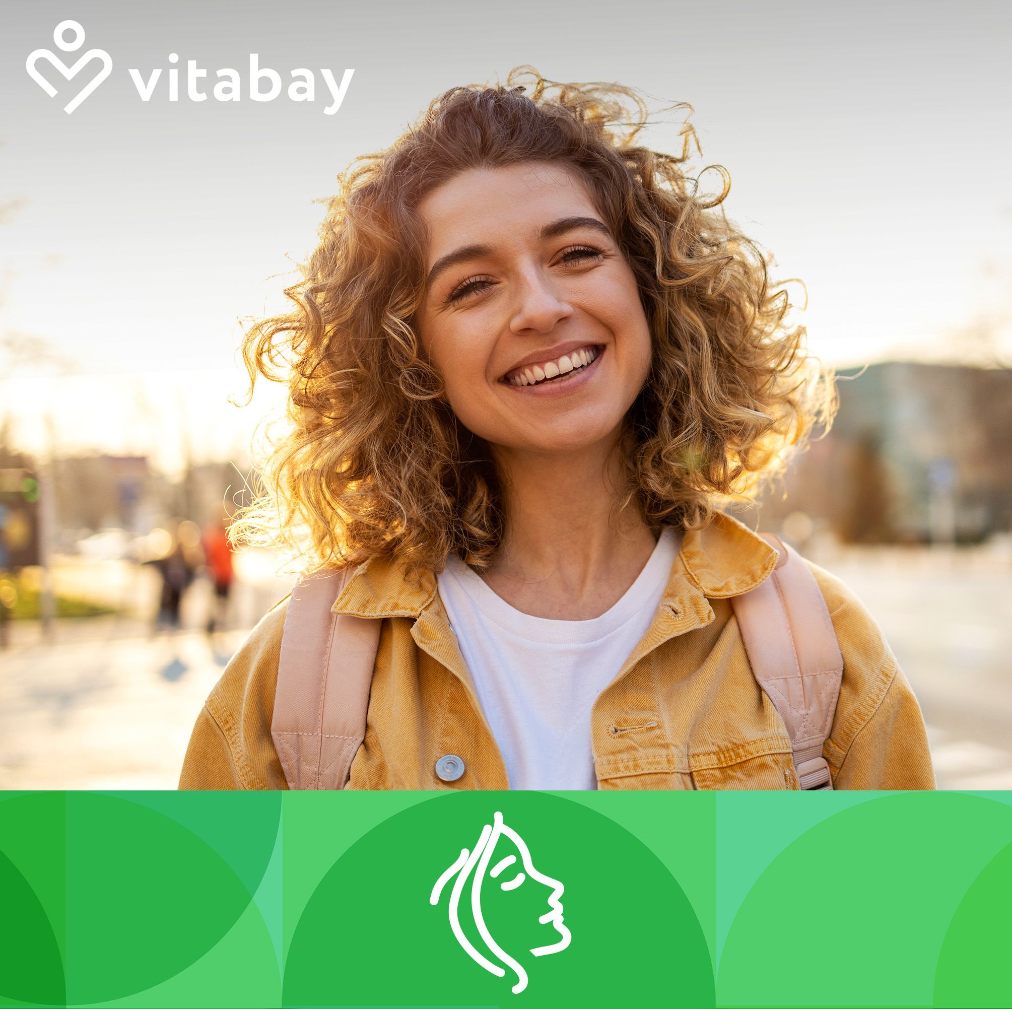 Eine lächelnde Frau mit lockigem Haar und Sonnenlicht im Hintergrund. Sie trägt eine gelbe Jacke und einen Rucksack.