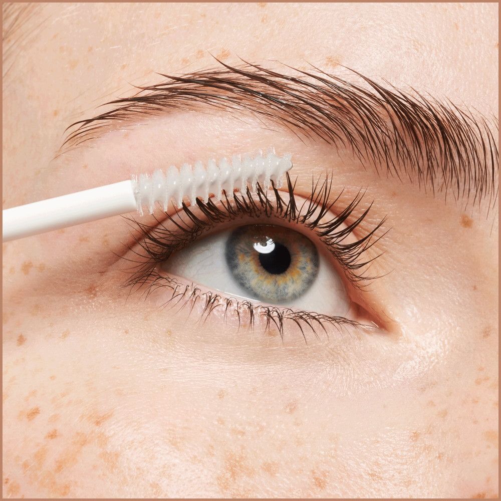 Auge mit Augenbraue. Bürste trägt Serum auf die Wimpern auf. Helle Haut mit Sommersprossen. Blaues Auge.